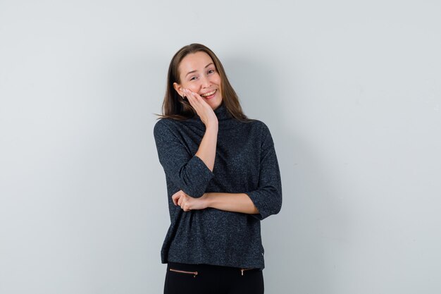 Young female posing while holding hand on her face in black blouse and looking jolly. front view.