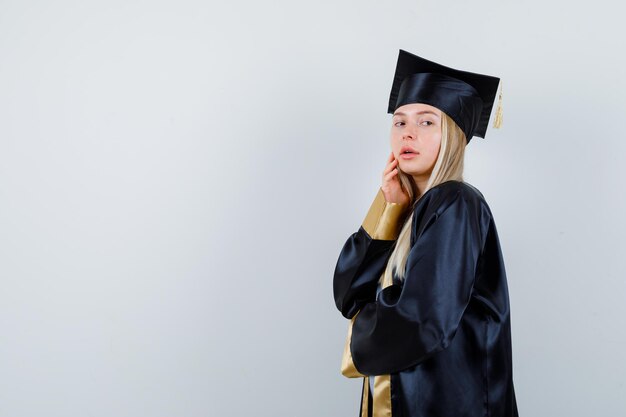 Young female posing while touching her cheek in graduate uniform and looking alluring. .