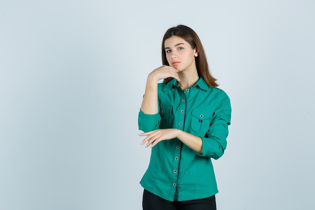 Young female posing while touching skin on her chin in green shirt and looking graceful , front view.