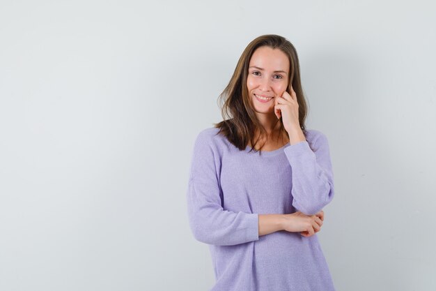 Young female posing with hand on cheek while smiling in lilac blouse and looking impressive 