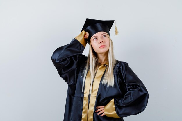 Young female posing with hand on head in graduate uniform and looking pensive