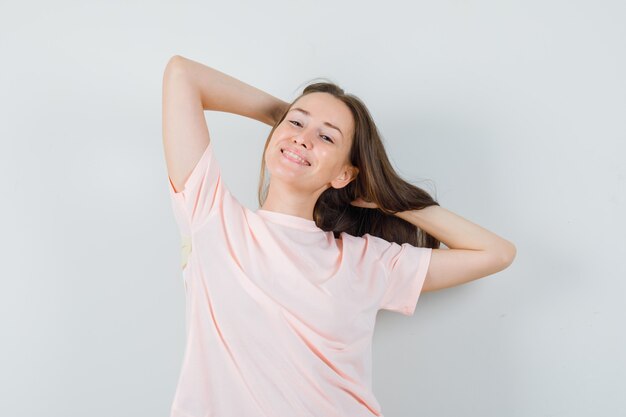 Молодая женщина позирует с руками в волосах в розовой футболке и выглядит очаровательно, вид спереди.