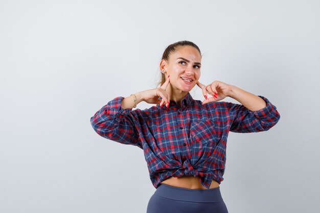 Young female pressing fingers on cheeks in checkered shirt, pants and looking cute , front view.