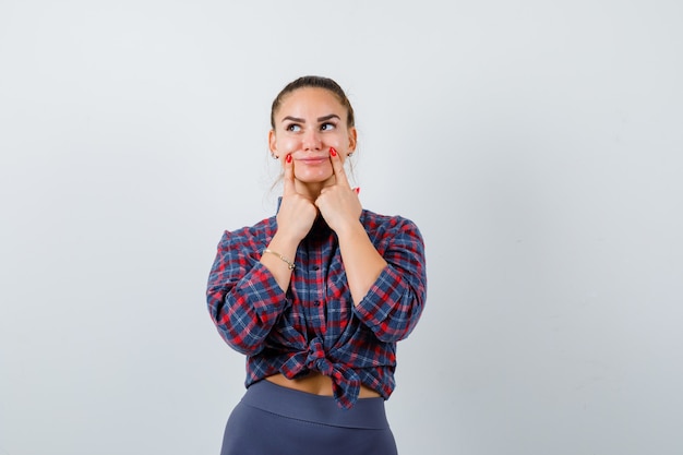 Giovane femmina premendo le dita sulle guance in camicia a scacchi, pantaloni e guardando pensieroso, vista frontale.