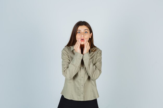 Young female pressing hands on cheeks in shirt, skirt and looking shocked