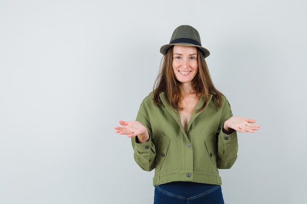 Foto gratuita giovane donna che finge di tenere o mostrare qualcosa in giacca, pantaloni, cappello e sembra felice. vista frontale.