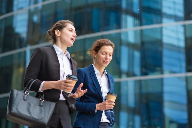 Young female professionals with takeaway coffee mugs wearing office suits, walking together past glass office building, talking, discussing project. Medium shot. Work break or friendship concept