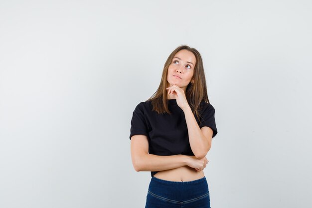 Young female propping chin on hand in black blouse, pants and looking dreamy