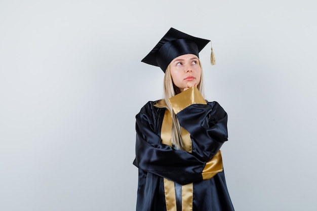 Young female propping chin on hand in graduate uniform and looking pensive