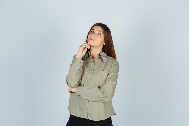 Young female propping chin on hand in shirt, skirt and looking pensive , front view.