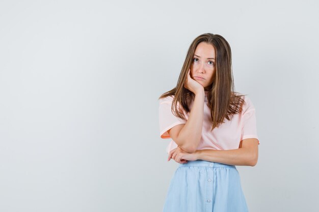 Young female propping chin on palm in t-shirt, skirt and looking hopeless , front view.