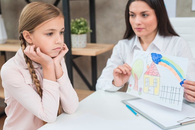 Free photo young female psychologist showing drawing paper with drawn house to sad serious girl