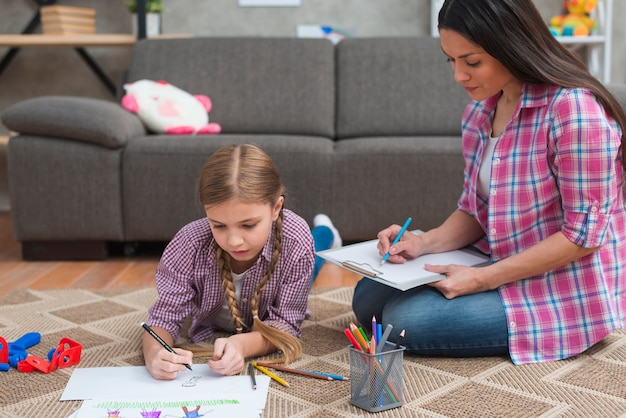 Young female psychologist taking notes while girl drawing on the white paper