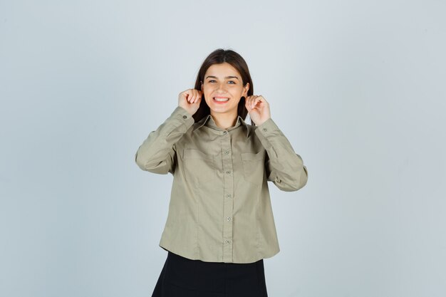 Young female pulling down her earlobes in shirt, skirt and looking merry