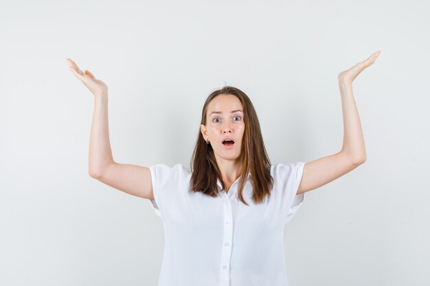 Young female raising arms for helpless gesture in white blouse and looking confused