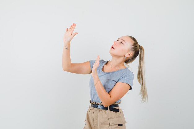 Young female raising hands to defend herself in t-shirt, pants and looking scared , front view.