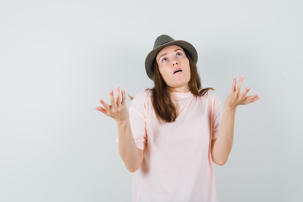 Young female raising hands in questioning manner in pink t-shirt, hat front view.