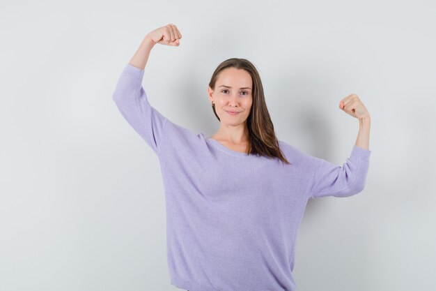 Young female raising her arms up while showing arm muscles in lilac blouse and looking powerful. front view.