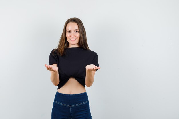 Young female raising open palms in black blouse, pants and looking cheerful.