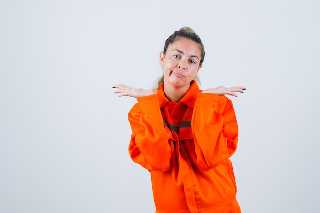 Young female raising up her open palms in worker uniform and looking puzzled. front view.