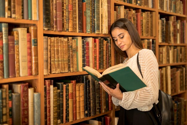 Young female reading book and leaning on shelf