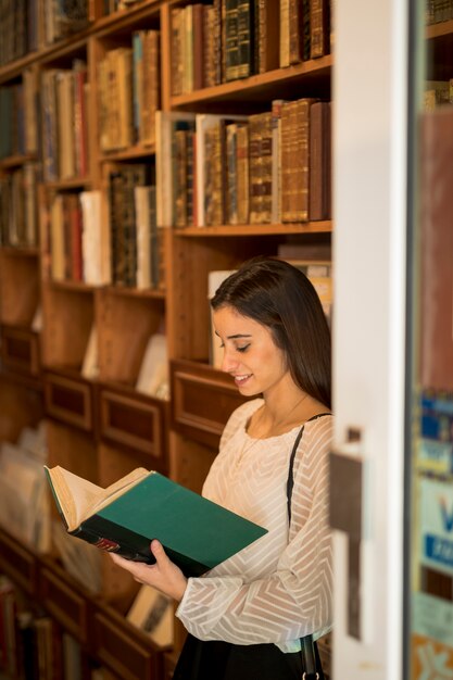 Молодая женщина, чтение книги возле книжной полки в библиотеке