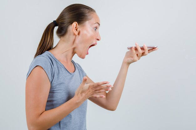 灰色のTシャツで叫び、怒っているように見えることによって音声メッセージを録音する若い女性。 。