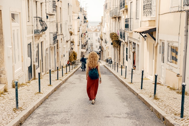 Free photo young female in a red dress walking through a road surrounded by buildings under the sunlight