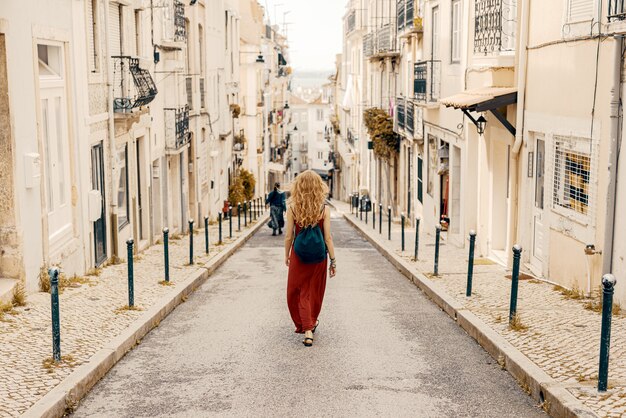 日光の下で建物に囲まれた道路を歩いている赤いドレスを着た若い女性