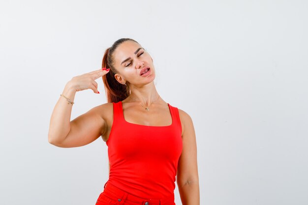 Young female in red tank top making suicide gesture and looking hopeless , front view.