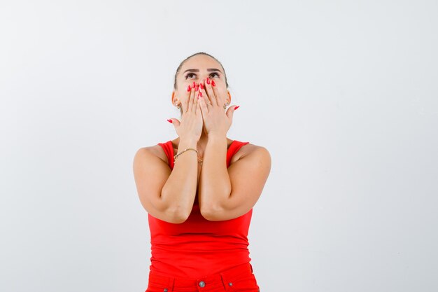 Giovane donna in canottiera rossa, pantaloni che tengono le mani sulla bocca mentre guarda in alto e guarda pensieroso, vista frontale.