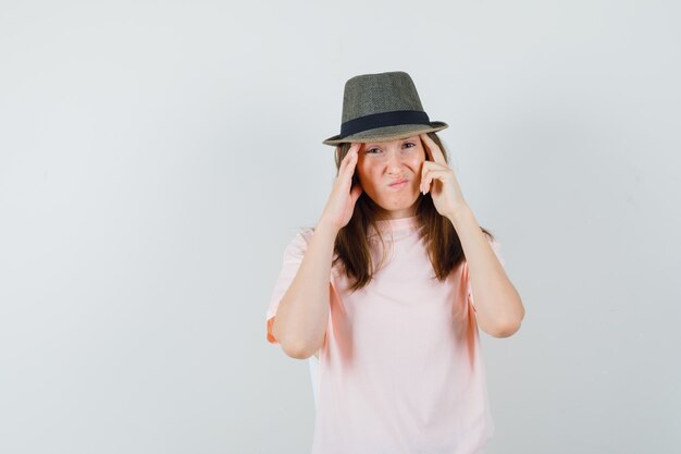 ピンクのTシャツ、帽子で彼女の寺院をこすり、動揺している若い女性。正面図。