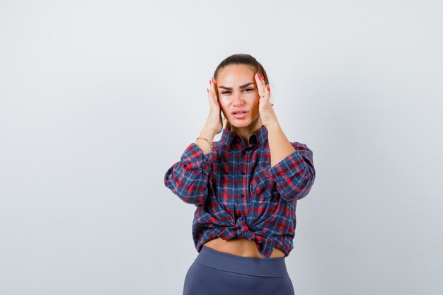 Young female rubbing temples in checkered shirt, pants and looking fatigued. front view.