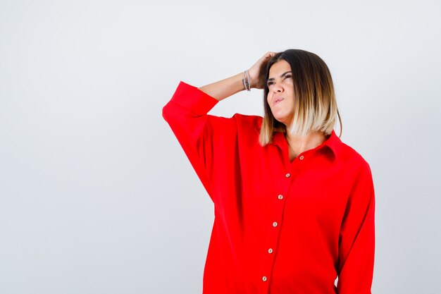 Young female scratching head while looking up in red oversized shirt and looking thoughtful , front view.