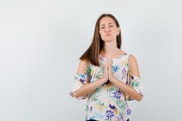 Young female in shirt holding hands in praying gesture and looking miserable , front view.