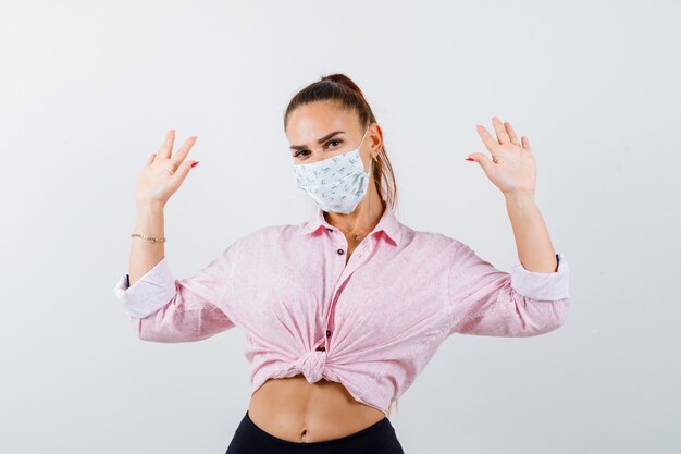 Young female in shirt, pants, medical mask showing palms in surrender gesture and looking serious , front view.