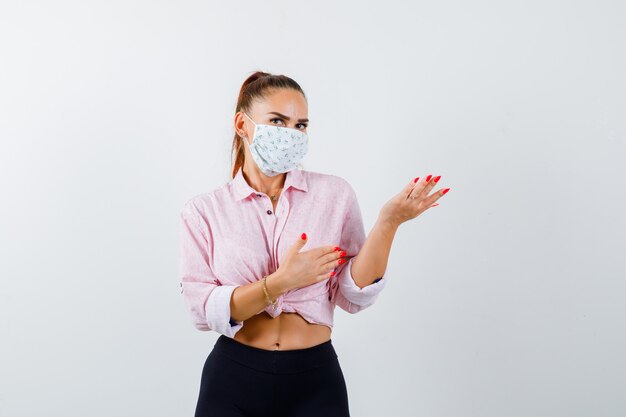 Young female in shirt, pants, medical mask welcoming something and looking confident , front view.