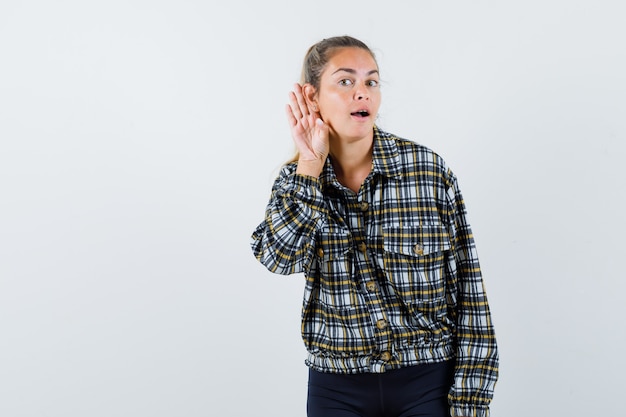 Young female in shirt, shorts holding hand behind ear and looking curious , front view.