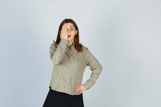 Young female in shirt, skirt holding hand on eye and looking wondered