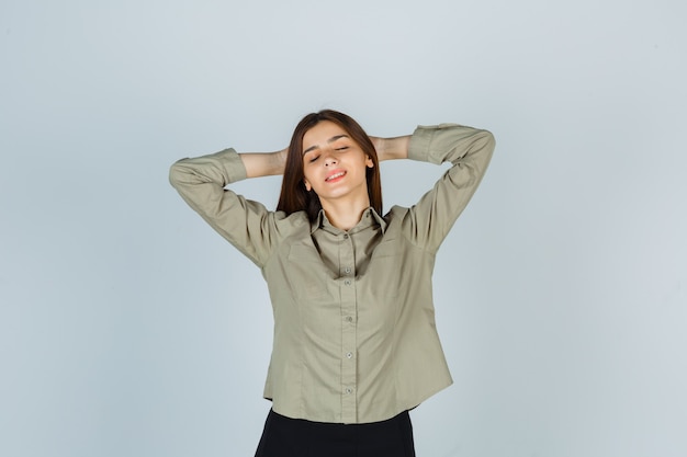 Foto gratuita giovane donna in camicia, gonna che tiene le mani dietro la testa e sembra rilassata, vista frontale.