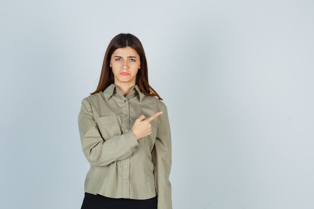 Young female in shirt, skirt pointing at upper right corner, curving lower lip and looking offended , front view.