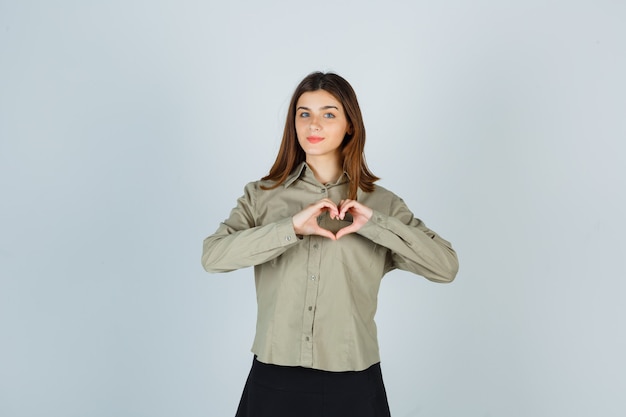 Foto gratuita giovane femmina in camicia, gonna che mostra il gesto del cuore e che sembra sicura