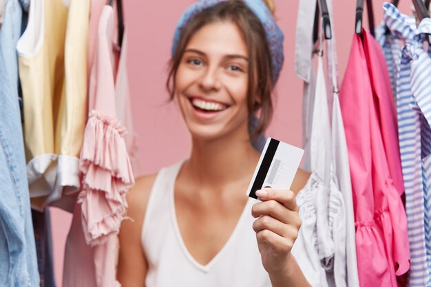 Young female shopaholic standing in dressing room near variety of clothes, holding plastic card in hands, going to buy new skirt