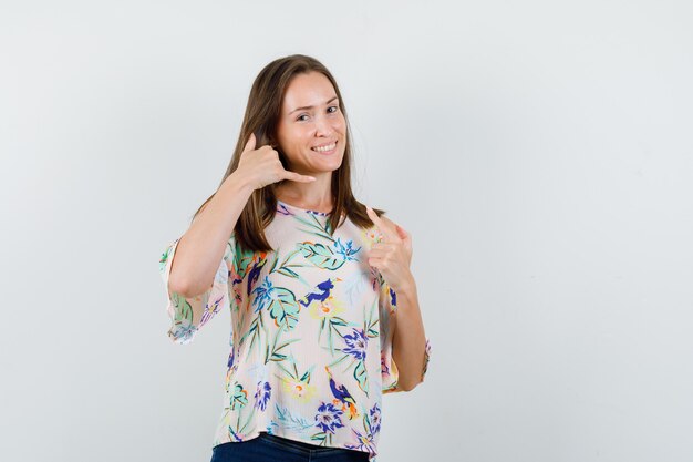Young female showing call me gesture in shirt, jeans and looking cheerful. front view.