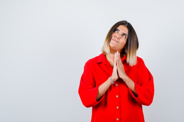 Young female showing clasped hands in pleading gesture in red oversized shirt and looking hopeful. front view.