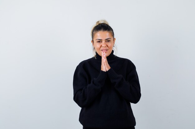 Young female showing clasped hands in pleading in turtleneck sweater and looking hopeful , front view.