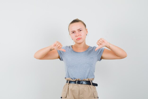 Молодая женщина показывает двойные пальцы вниз в футболке, брюках и выглядит разочарованной, вид спереди.