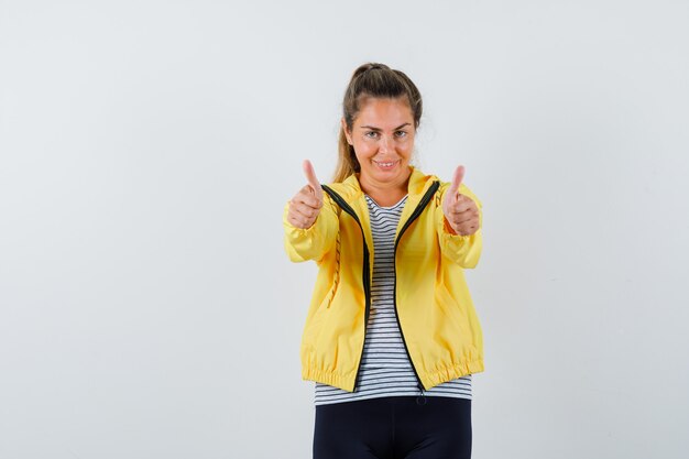 Young female showing double thumbs up in jacket, t-shirt and looking happy. front view.