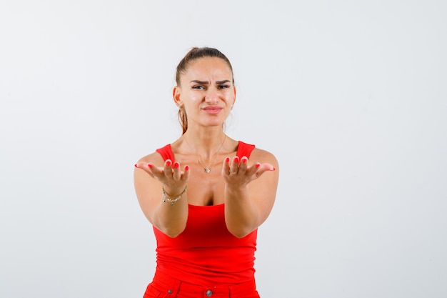 Young female showing giving gesture in red tank top, pants and looking cute , front view.