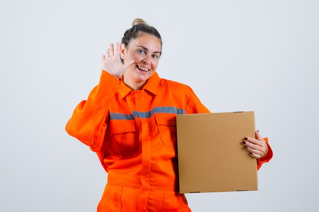 Young female showing goodbye gesture while holding box in worker uniform and looking glad. front view.